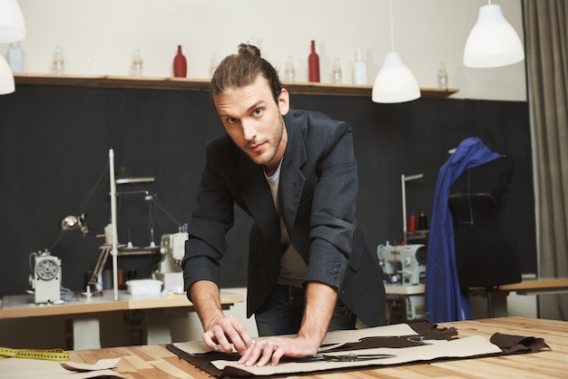 Retrato del talentoso diseñador masculino joven y guapo con elegante peinado y atuendo informal mirando a la cámara con expresión de la cara concentrada mientras recorta partes del vestido de la colección de primavera