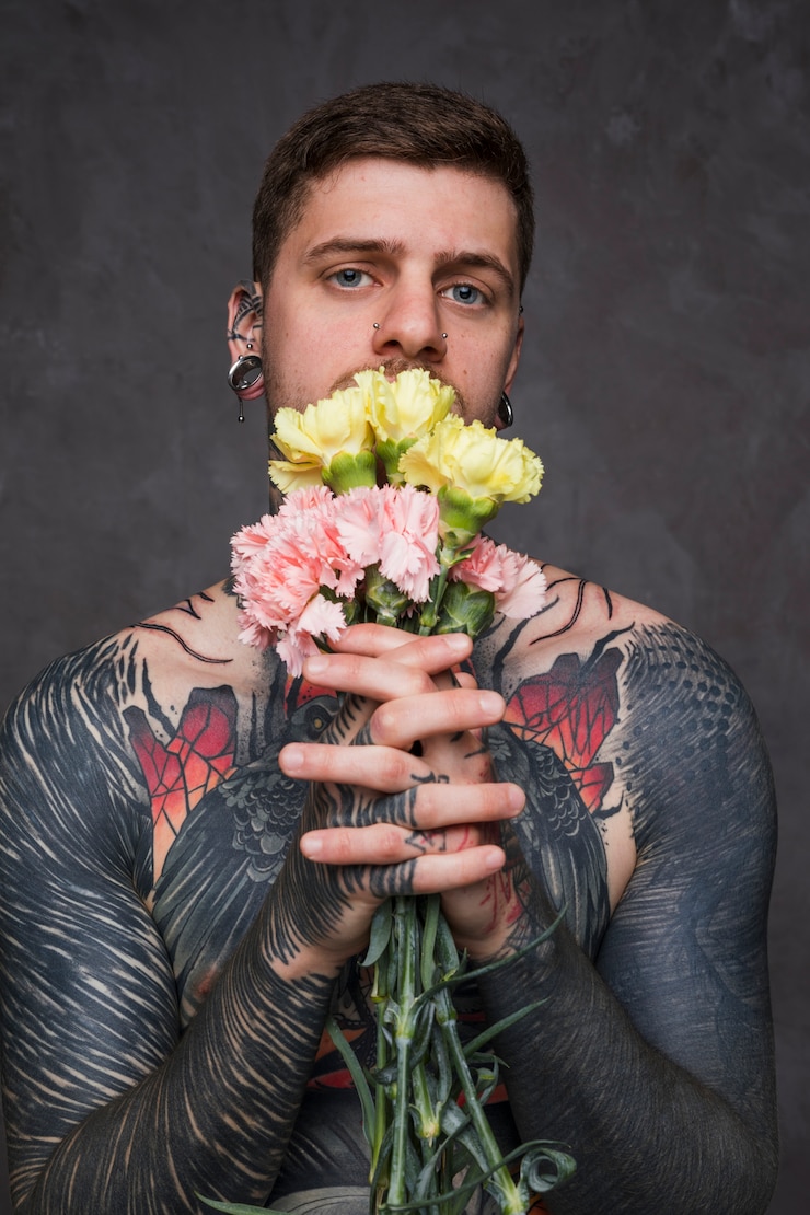 Foto gratuito retrato de un tatuaje y un hombre joven perforado que sostiene la flor del clavel en manos unidas