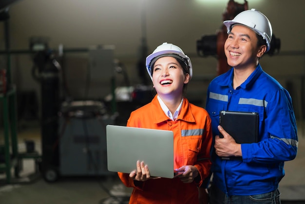 Retrato de una técnica asiática emgineer masculina con uniforme de seguridad de pie y dando la vuelta para mirar la cámara y reír con una sonrisa alegre y confiada en el fondo del lugar de trabajo de la fábrica de maquinaria