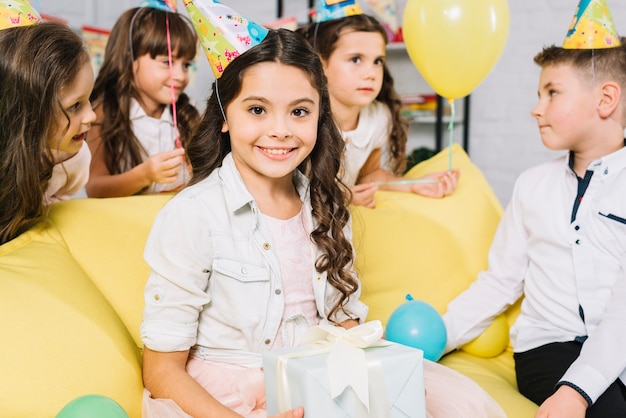 El retrato de una tenencia de la muchacha del feliz cumpleaños presenta en la mano que se sienta en el sofá con sus amigos