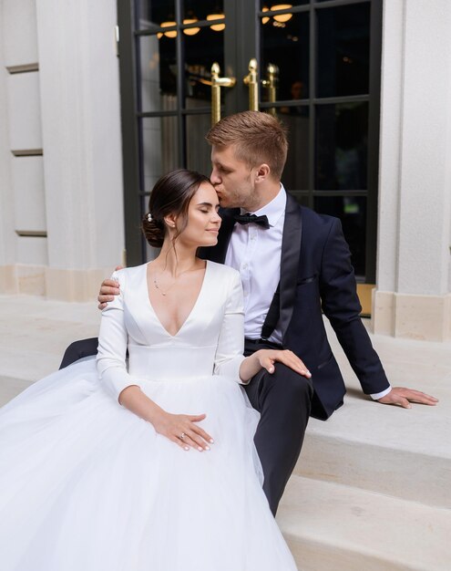 Retrato de ternura pareja casada novio besando a la esposa en la frente con un hermoso vestido blanco Pareja amorosa sentada en las escaleras disfrutando de la ceremonia del día de la boda Matrimonio Familia