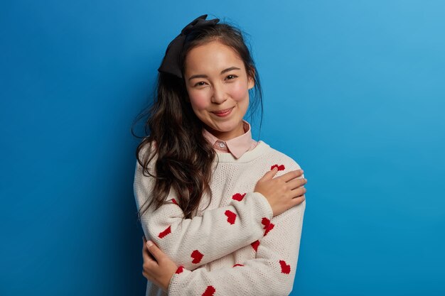 Retrato de tierna joven asiática siente comodidad y tranquilidad, se abraza a ella, sonríe agradablemente, vestida con un jersey suave expresa emociones positivas aisladas en la pared azul cruza los brazos sobre el cuerpo