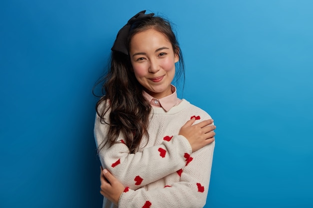 Retrato de tierna joven asiática siente comodidad y tranquilidad, se abraza a ella, sonríe agradablemente, vestida con un jersey suave expresa emociones positivas aisladas en la pared azul cruza los brazos sobre el cuerpo