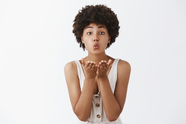 Retrato de tierna y linda mujer afroamericana encantadora con peinado rizado, doblando los labios y sosteniendo las palmas cerca de la boca para enviar un beso de viento sobre la pared gris