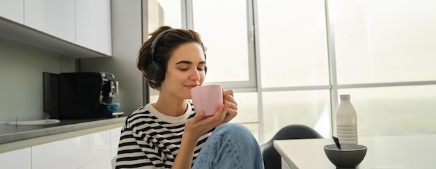 Retrato de tierna mujer hermosa con té escucha música en los auriculares sostiene la taza y sonríe