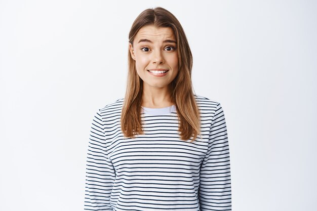 Retrato de tímida chica rubia nerviosa mordiéndose el labio y sonriendo incómodo, sintiéndose inseguro, de pie emocionado contra la pared blanca