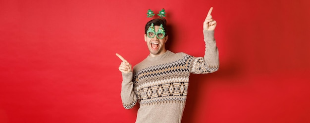 Retrato de un tipo alegre y apuesto con gafas de fiesta y suéter navideño, bailando y señalando con el dedo a los lados, disfrutando de la celebración del año nuevo, de pie sobre un fondo rojo.