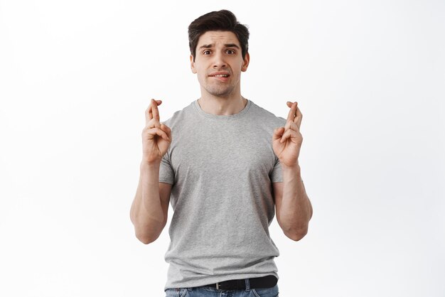 Retrato de un tipo nervioso que cruza los dedos para tener suerte mordiéndose el labio y frunciendo el ceño preocupado esperando noticias importantes anticipando resultados deseando pararse sobre fondo blanco