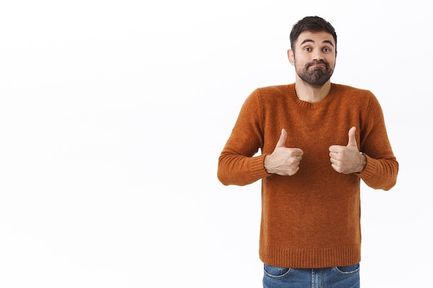 Foto gratuita retrato de tonto y guapo chico normal sonriente, hombre barbudo que muestra el pulgar hacia arriba y se encoge de hombros, la sonrisa dice que no está mal, anima a un amigo con un resultado normal promedio, pared blanca