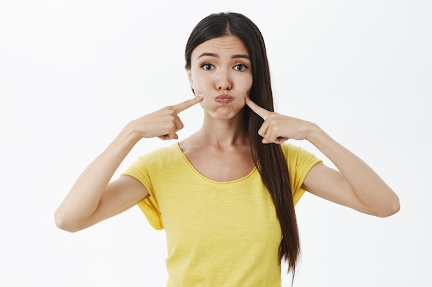 Retrato de tonto modelo femenino femenino y lindo en camiseta amarilla haciendo pucheros conteniendo la respiración y tocando las mejillas con los dedos índices