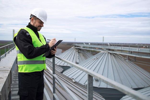 Foto gratuita retrato de trabajador de la construcción de pie sobre los tejados de tanques de almacenamiento de silos altos y trabajando en una tableta