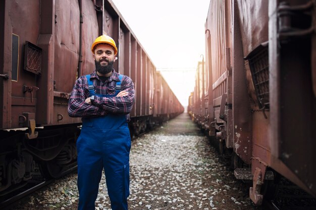 Retrato de trabajador del ferrocarril con los brazos cruzados de pie con orgullo en la estación de tren entre vagones