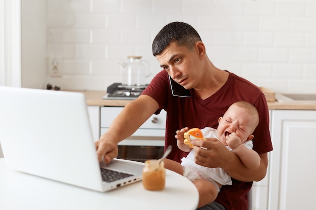 Retrato de trabajador independiente masculino morena ocupado con camiseta marrón estilo casual sentado a la mesa en la cocina con su hija pequeña y hablando por teléfono celular con el cliente.