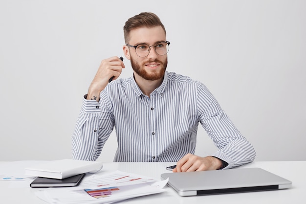 Retrato de trabajador de oficina masculino, tiene apariencia específica, vestido formalmente, se sienta en el escritorio blanco,
