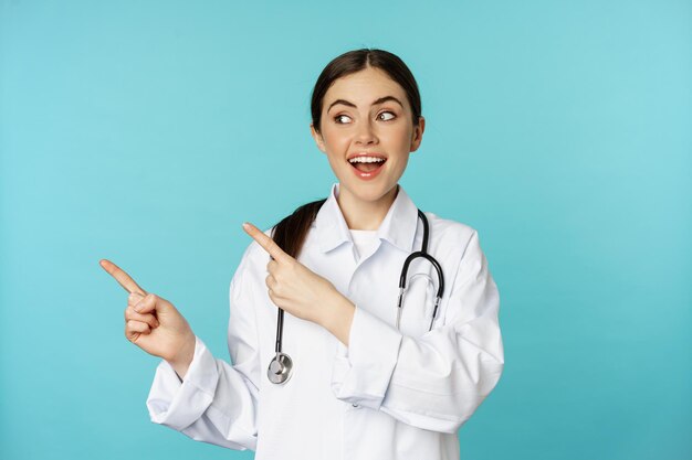 Retrato de una trabajadora médica sonriente, una doctora con bata blanca con estetoscopio, señalando con el dedo a la izquierda, mostrando un anuncio de clínica médica, fondo turquesa