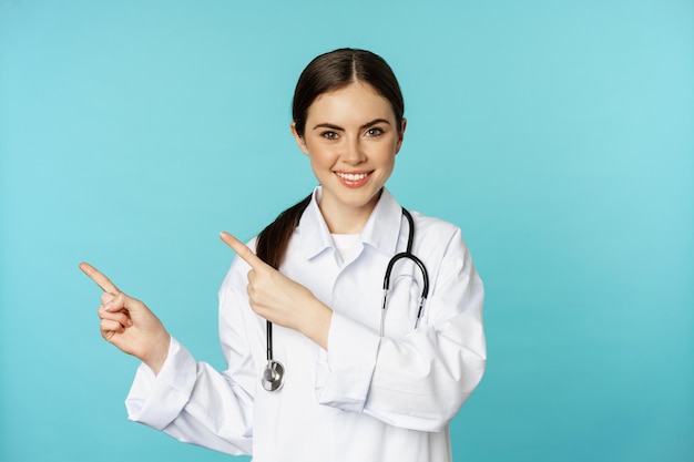 Retrato de una trabajadora médica sonriente, una doctora con bata blanca con estetoscopio, señalando con el dedo a la izquierda, mostrando un anuncio de clínica médica, fondo turquesa