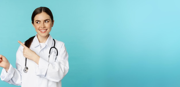 Foto gratuita retrato de una trabajadora médica sonriente doctora en bata blanca con estetoscopio señalando con el dedo a la izquierda mostrando el fondo turquesa del anuncio de la clínica médica