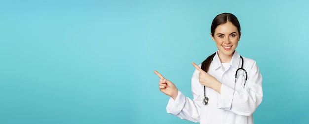 Foto gratuita retrato de una trabajadora médica sonriente doctora en bata blanca con estetoscopio señalando con el dedo a la izquierda