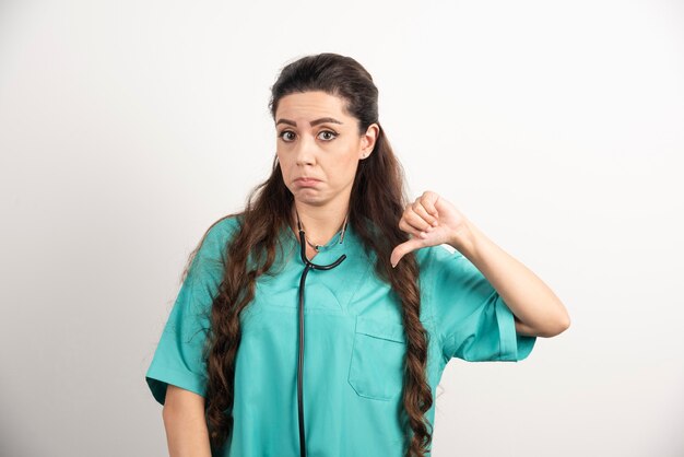 Retrato de trabajadora sanitaria posando con el pulgar hacia abajo en la pared blanca.