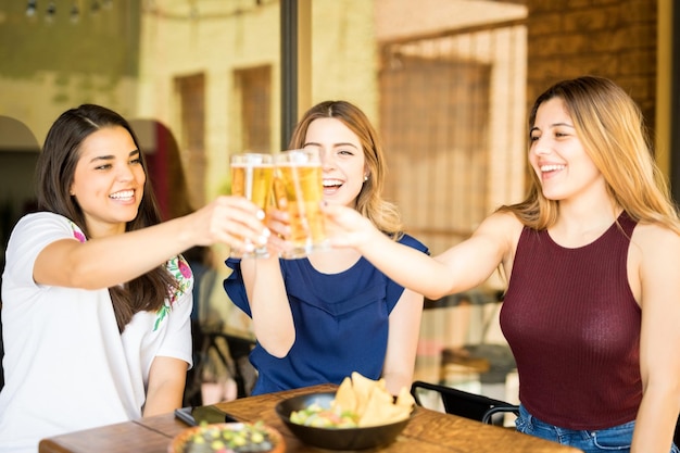 Retrato de tres amigas jóvenes tomando cervezas en el café, brindando vasos de cerveza y sonriendo.
