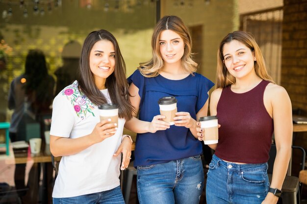 Retrato de tres amigas de pie afuera de un restaurante con una taza de café y haciendo contacto visual
