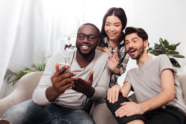 Retrato de tres amigos sonrientes sentados en sillas en casa y felizmente mirando en el teléfono móvil mientras pasan tiempo juntos aislados