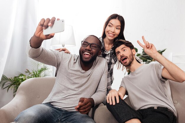 Retrato de tres amigos sonrientes sentados en sillas en casa y tomando fotos divertidas en la cámara frontal del teléfono móvil juntos aislados