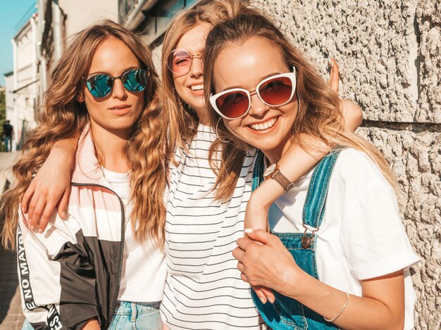 Retrato de tres jóvenes hermosas chicas hipster sonrientes en ropa de moda de verano. Mujeres despreocupadas sexy posando en la calle cerca de la pared. Modelos positivos divirtiéndose en gafas de sol.