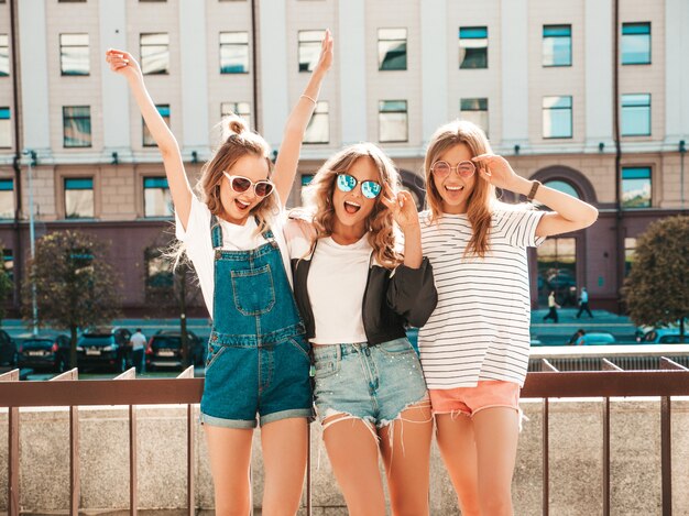 Retrato de tres jóvenes hermosas chicas hipster sonrientes en ropa de moda de verano. Mujeres despreocupadas sexy posando en la calle. Modelos positivos divirtiéndose en gafas de sol. Levantando las manos