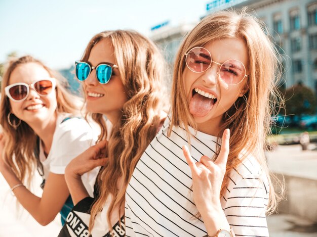 Retrato de tres jóvenes hermosas chicas hipster sonrientes en ropa de moda de verano. Mujeres despreocupadas sexy posando en la calle. Modelos positivos divirtiéndose en gafas de sol. Muestra el signo de rock and roll