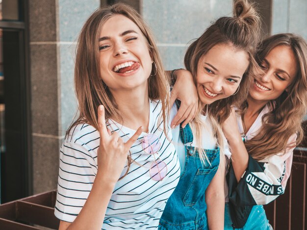 Retrato de tres jóvenes hermosas chicas hipster sonrientes en ropa de moda de verano. Mujeres despreocupadas y sexy posando en la calle. Modelos positivos divirtiéndose. Muestran lengua y rock and roll.