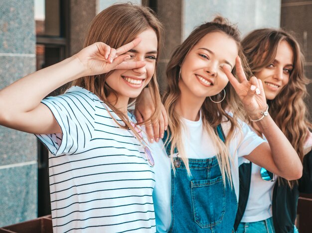 Retrato de tres jóvenes hermosas chicas hipster sonrientes en ropa de moda de verano. Mujeres despreocupadas y sexy posando en la calle.Los modelos positivos muestran signos de paz