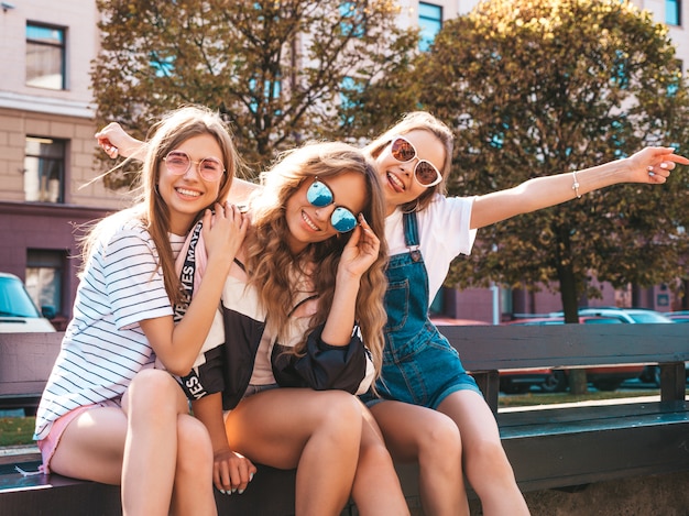 Retrato de tres jóvenes hermosas chicas hipster sonrientes en ropa de moda de verano. Mujeres despreocupadas y sexys sentadas en el banco en la calle. Modelos positivos divirtiéndose en gafas de sol. Levantando las manos