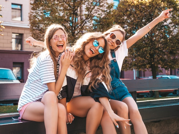 Retrato de tres jóvenes hermosas chicas hipster sonrientes en ropa de moda de verano. Mujeres despreocupadas y sexys sentadas en el banco en la calle. Modelos positivos divirtiéndose en gafas de sol. Levantando las manos