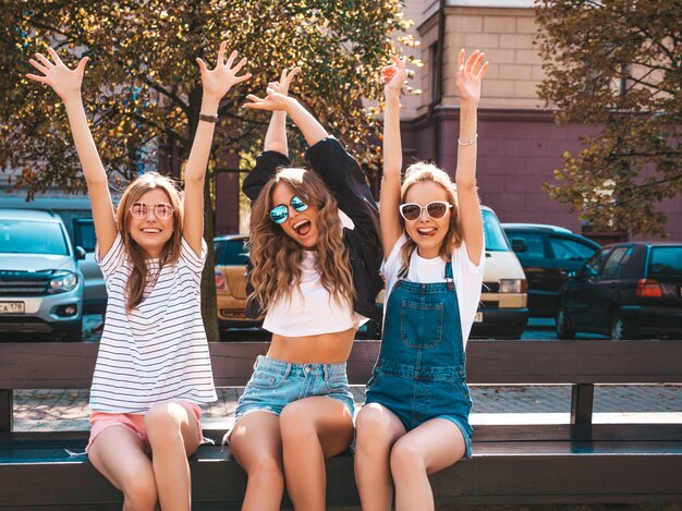 Retrato de tres jóvenes hermosas chicas hipster sonrientes en ropa de moda de verano. Mujeres despreocupadas y sexys sentadas en el banco en la calle. Modelos positivos divirtiéndose en gafas de sol. Levantando las manos