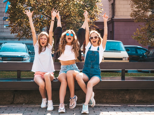 Retrato de tres jóvenes hermosas chicas hipster sonrientes en ropa de moda de verano. Mujeres despreocupadas y sexys sentadas en el banco en la calle. Modelos positivos divirtiéndose en gafas de sol. Levantando las manos