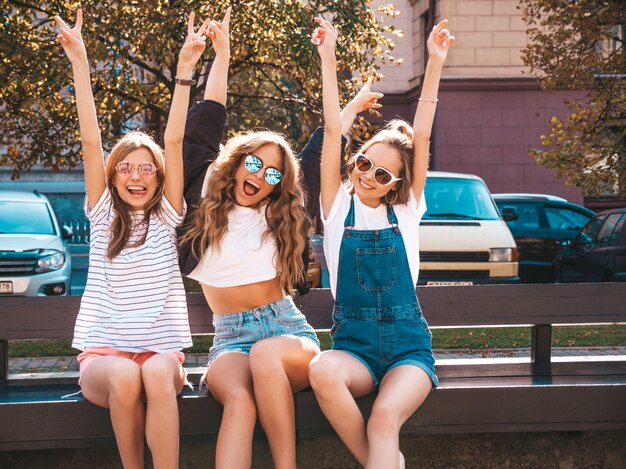 Retrato de tres jóvenes hermosas chicas hipster sonrientes en ropa de moda de verano. Mujeres despreocupadas y sexys sentadas en el banco en la calle. Modelos positivos divirtiéndose en gafas de sol. Levantando las manos