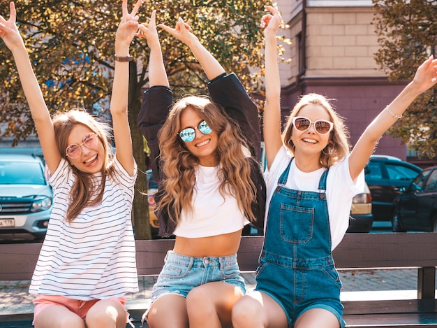 Retrato de tres jóvenes hermosas chicas hipster sonrientes en ropa de moda de verano. Mujeres despreocupadas y sexys sentadas en el banco en la calle. Modelos positivos divirtiéndose en gafas de sol. Levantando las manos