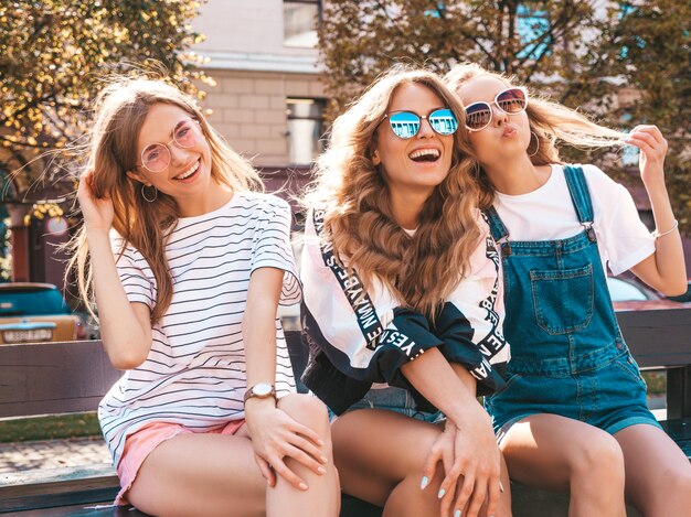 Retrato de tres jóvenes hermosas chicas hipster sonrientes en ropa de moda de verano. Mujeres despreocupadas sexys sentadas en el banco en la calle. Modelos positivos divirtiéndose en gafas de sol