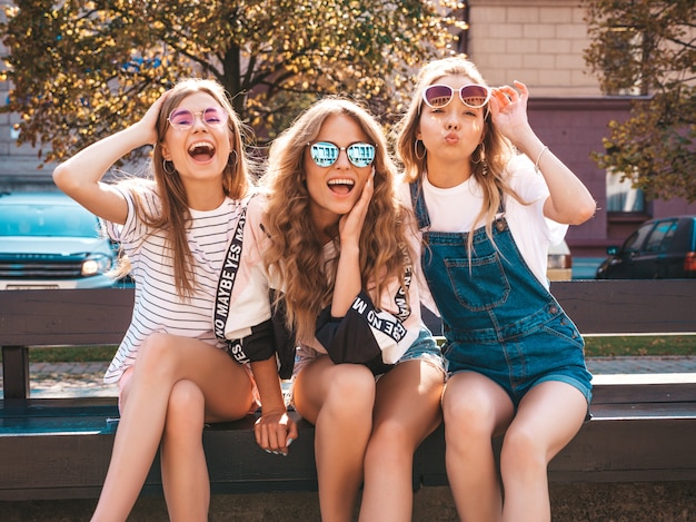 Retrato de tres jóvenes hermosas chicas hipster sonrientes en ropa de moda de verano. Mujeres despreocupadas sexys sentadas en el banco en la calle. Modelos positivos divirtiéndose en gafas de sol