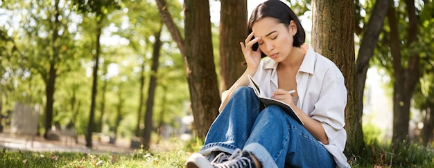 Foto gratuita retrato de una triste chica asiática escribiendo en su diario y sintiéndose incómoda sentada sola en el parque bajo un árbol