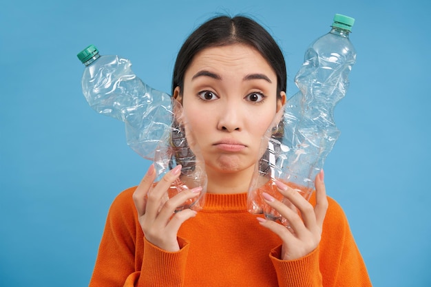 Retrato de una triste mujer asiática con botellas de plástico molesta por la falta de centros de reciclaje de fondo azul.