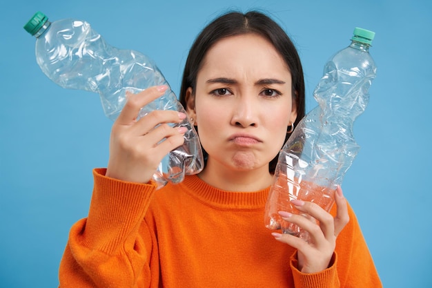 Foto gratuita retrato de una triste mujer asiática con botellas de plástico molesta por la falta de centros de reciclaje de fondo azul.