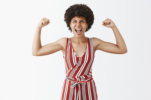 Retrato de triunfante deportista afroamericana guapa alegre y expresiva con elegantes monos a rayas que levantan los brazos para mostrar los músculos gritando de alegría y mirando hacia arriba