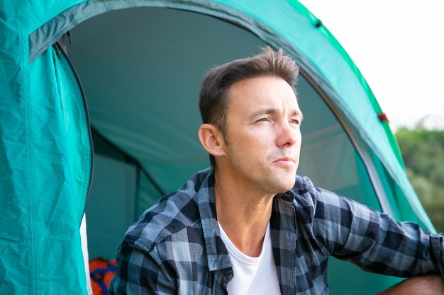 Retrato de turista masculino sentado en la tienda y mirando a otro lado. Jóvenes excursionistas caucásicos o viajero relajándose en la naturaleza y disfrutando del paisaje. Turismo de mochilero, aventura y concepto de vacaciones de verano.