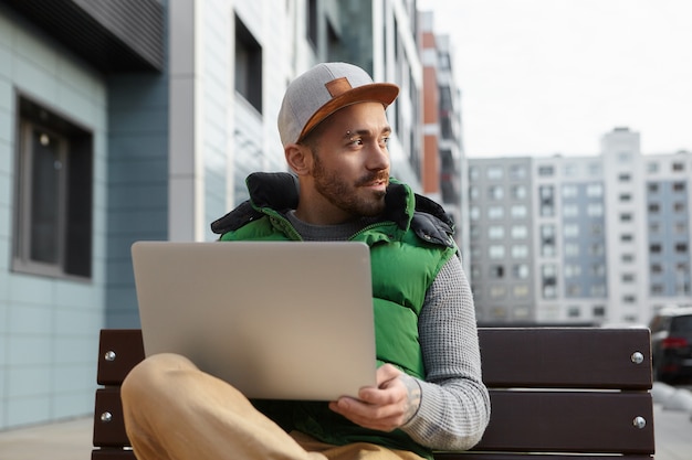 Retrato urbano de guapo joven europeo rastrojo trabajando de forma remota sentado en un banco con una computadora portátil en su regazo contra edificios borrosos, mirando hacia los lados y sonriendo