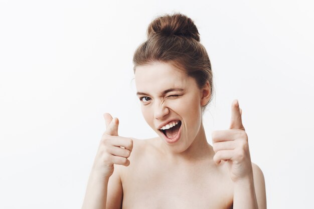 Retrato de ute chica caucásica guapa divertida con cabello largo y oscuro en peinado de moño y hombros desnudos sonriendo con dientes, guiñando un ojo con expresión feliz y coqueta