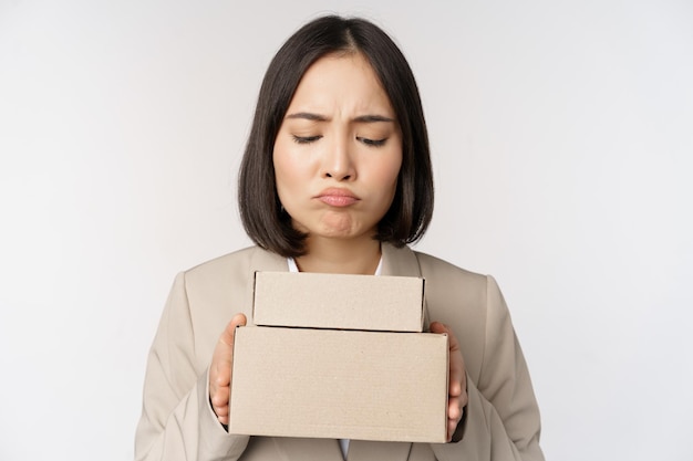 Retrato de una vendedora asiática empresaria sosteniendo cajas y luciendo triste y decepcionada de pie sobre fondo blanco