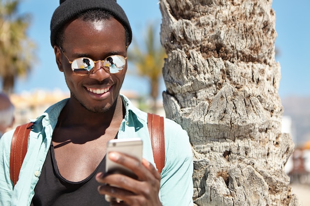 Retrato de verano al aire libre del alegre chico de piel oscura con ropa de moda usando un teléfono celular, disfrutando de la comunicación en línea con amigos a través de redes sociales, mensajes, enviando fotos durante un viaje al extranjero