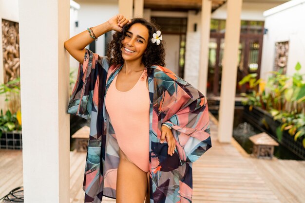 Retrato de verano al aire libre de una linda mujer morena con flores tropicales en los pelos posando en un hotel elegante